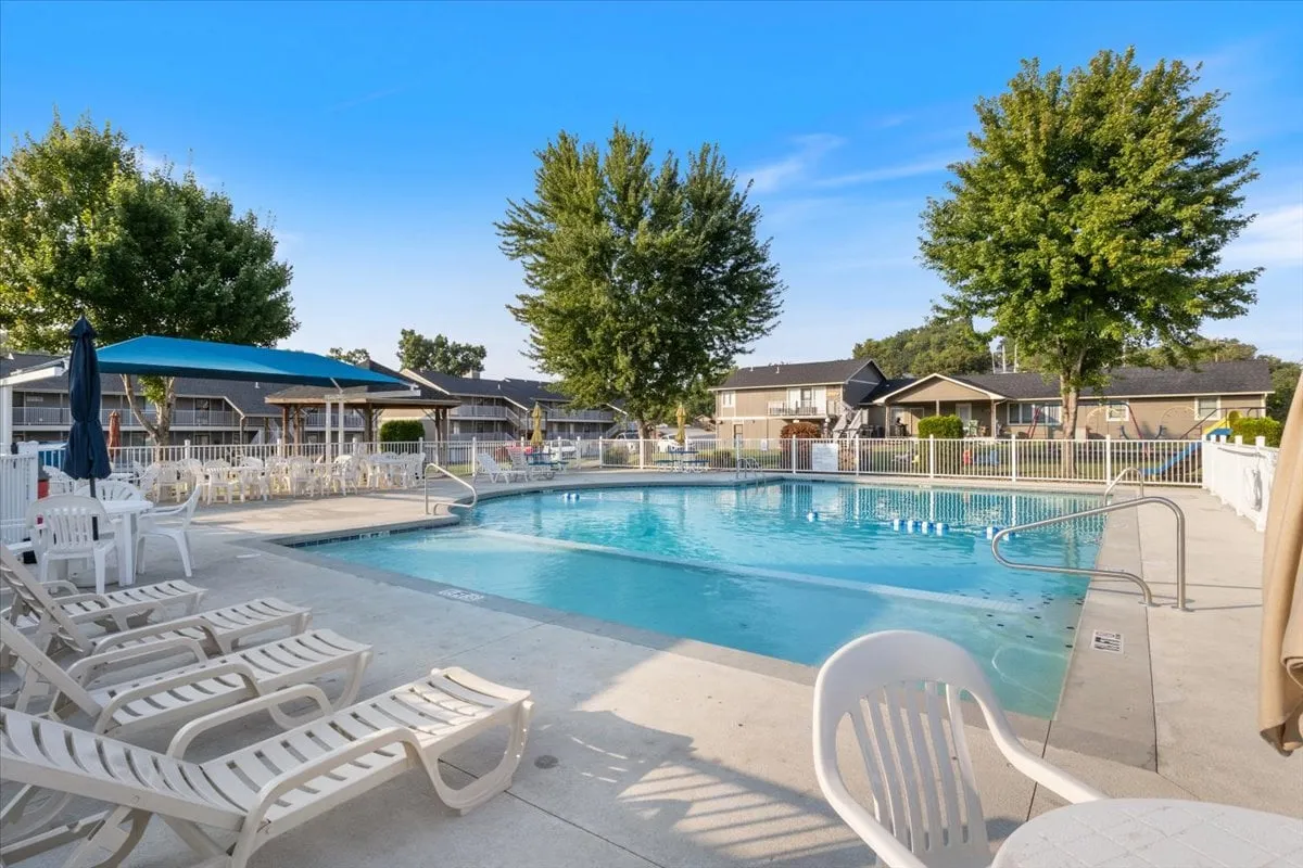 This image features a sparkling outdoor swimming pool surrounded by lush trees, with several lounge chairs and poolside umbrellas. The inviting pool area looks ready for kids and families to enjoy, suggesting summer fun and relaxation just steps from home.