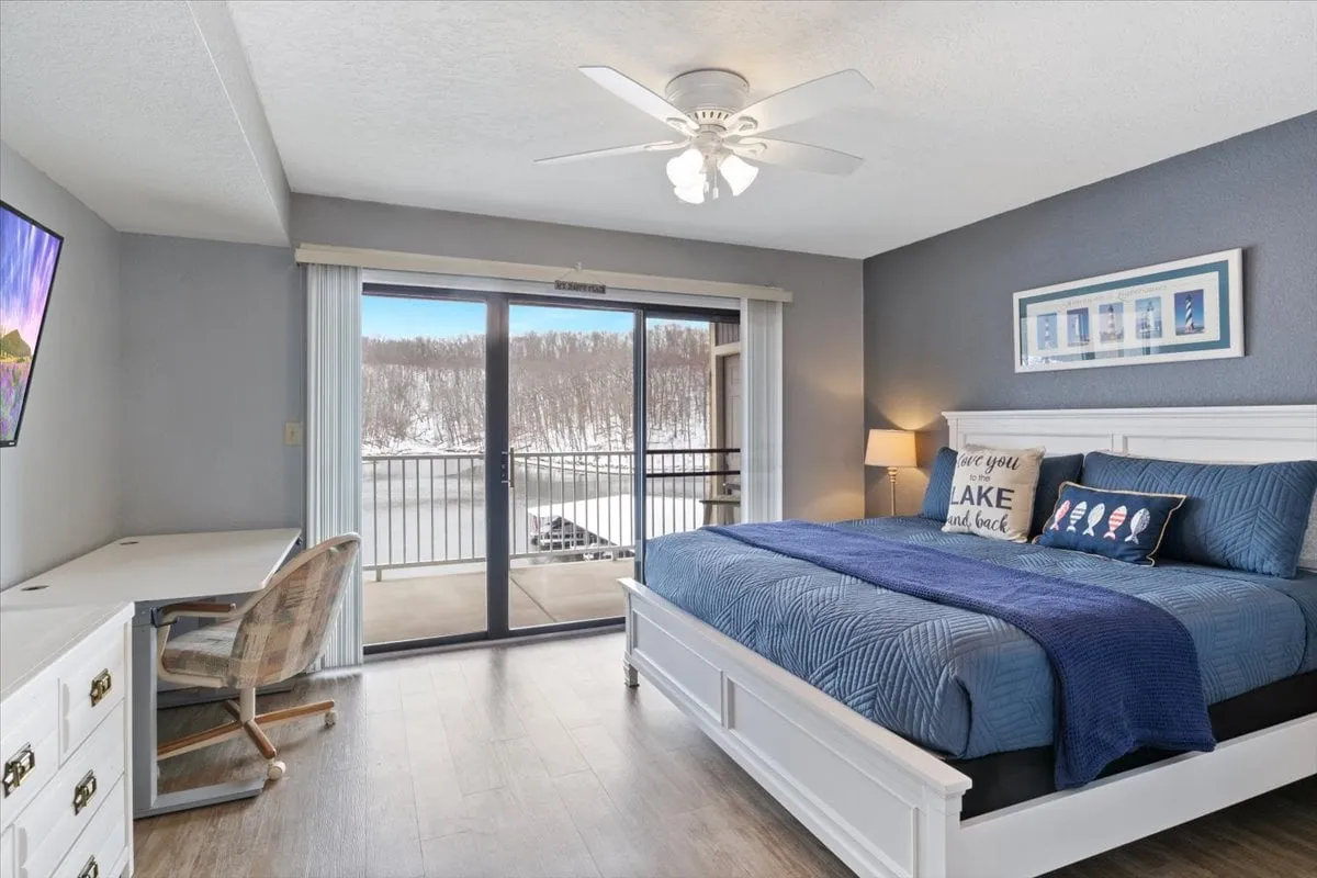 This image features a serene bedroom with a comfortable bed, a balcony view overlooking the lake, a dedicated desk for working, a television, and a warm lamp. The calming color palette and soft lighting create an inviting and private atmosphere ideal for both relaxation and productivity.