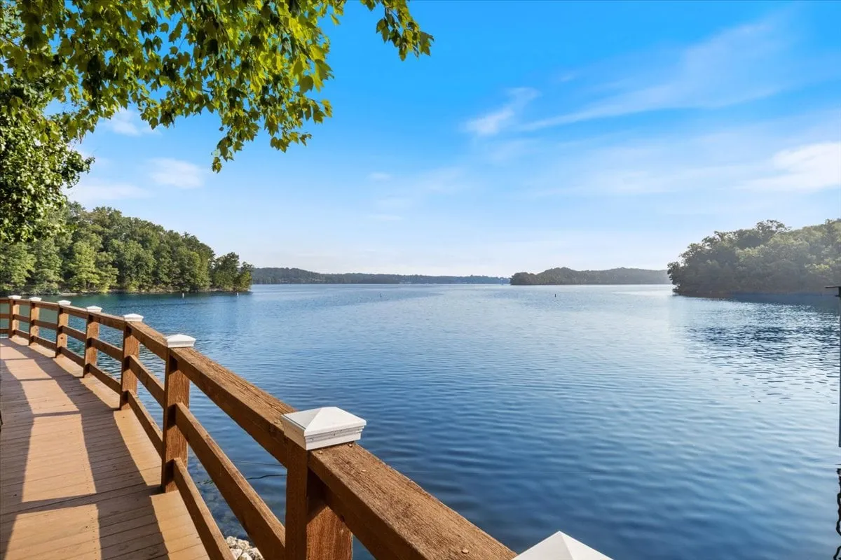 The hero image showcases a tranquil view of the Lake of the Ozarks from the condo's wooden-railed balcony, with calm water gently reflecting the tree-lined shore. The scene captures the serenity of lakeside living and invites you to imagine sunset strolls, fishing, or simply relaxing while soaking in the peaceful ambiance.