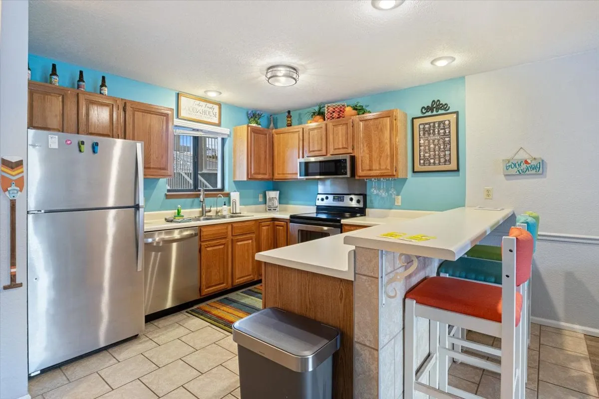 A bright, modern kitchen with stainless steel appliances, a large refrigerator, colorful bar stools at a kitchen island, and decorative wall art. The space feels spacious, practical, and welcoming—inviting for both big family meals and quick snacks.
