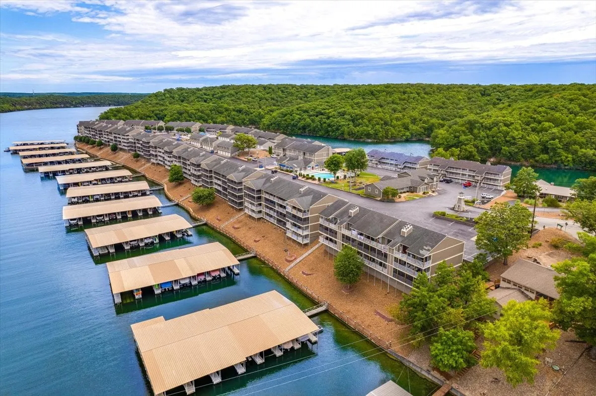A vibrant waterfront scene shows boathouses, landscaped areas, and multi-story buildings right on the water’s edge. Families can be imagined fishing, swimming, or simply relaxing by the lake. The layout highlights proximity to the water, boat access, and places to launch kayaks or paddle boards.