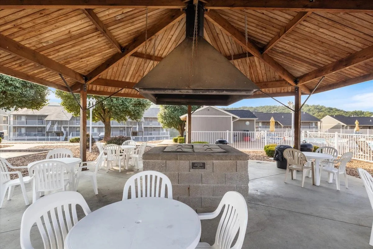 An exterior view of a pavilion with a grill, tables, and chairs nestled under the wooden structure. People are gathered around, enjoying a barbecue, with the atmosphere exuding casual group fun and togetherness.